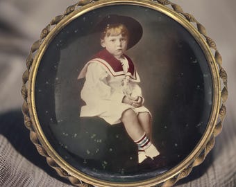 May include: A vintage framed photograph of a young boy wearing a sailor outfit and a hat. The photo is in a gold-coloured frame with a rope-like design.