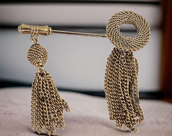 Op de afbeelding: Een gouden broche met twee hangende kwastjes. De kwastjes zijn gemaakt van gekoppelde kettingen en de bovenkant van de broche is een cirkel van gekoppelde kettingen.