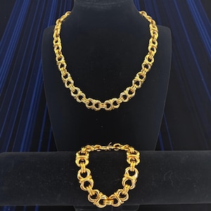Puede incluir: Un conjunto de collar y pulsera dorados. El collar se exhibe sobre un busto negro y la pulsera sobre un soporte negro. Ambas piezas presentan un diseño de eslabones repetidos. Las joyas se presentan sobre un fondo azul oscuro.