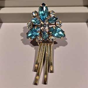 May include: A vintage brooch with a floral design. The brooch features large, light blue, marquise-cut rhinestones and smaller, clear, round rhinestones set in a gold-tone metal. Three gold-tone bars dangle from the bottom of the brooch.