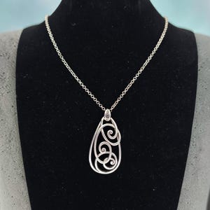 Może przedstawiać: Srebrny naszyjnik z wisiorkiem w kształcie łzy. Wisiorek ma ażurowy wz&oacute;r z wirującymi wzorami. Naszyjnik jest prezentowany na czarnej powierzchni.