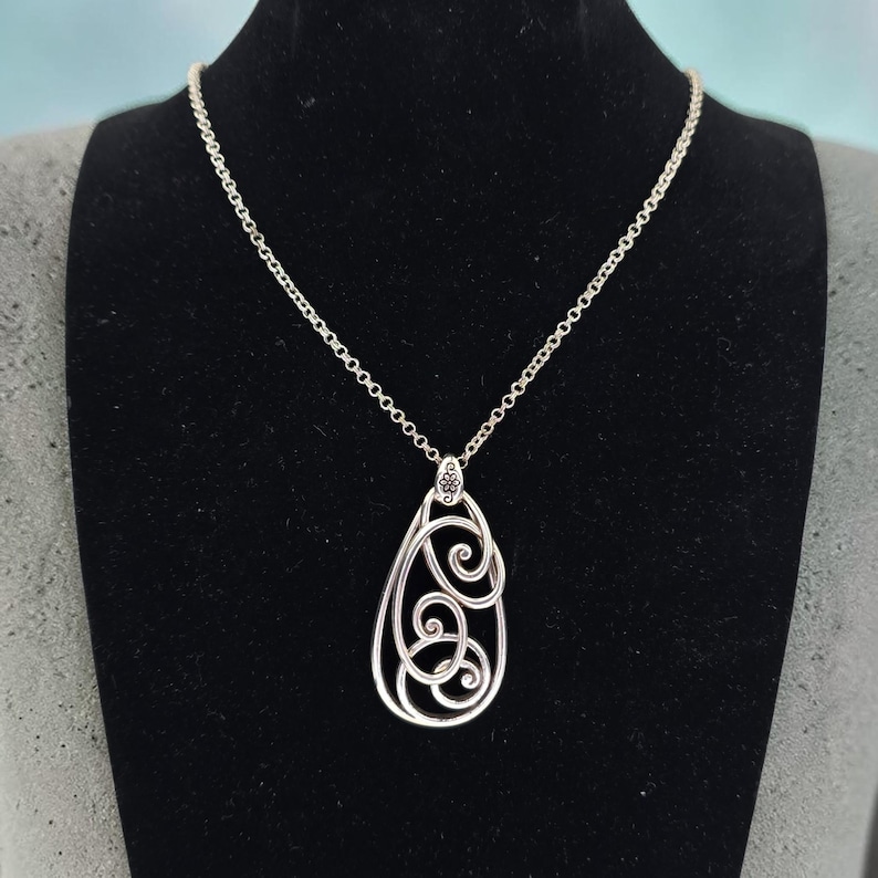 Może przedstawiać: Srebrny naszyjnik z wisiorkiem w kształcie łzy. Wisiorek ma ażurowy wz&oacute;r z wirującymi wzorami. Naszyjnik jest prezentowany na czarnej powierzchni.