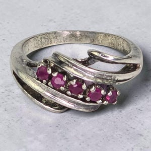 May include: A silver-toned ring featuring a bypass-style band with a polished finish. The center of the ring is set with five small, round, deep pinkish-red gemstones arranged in a single horizontal row, nestled between the sweeping metal curves.