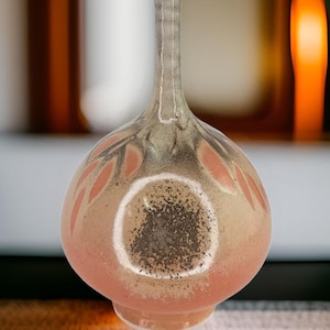 Puede incluir: Un jarrón pequeño, redondo y de cerámica con cuello estrecho. El jarrón está decorado con un esmalte rosa claro y gris y tiene un sutil patrón de hojas.