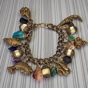 Può includere: Un braccialetto con ciondoli in stile vintage con una catena in bronzo e vari ciondoli. I ciondoli includono un pesce, una teiera e pietre preziose nei toni del blu, verde e viola. Il braccialetto ha una chiusura a moschettone.
