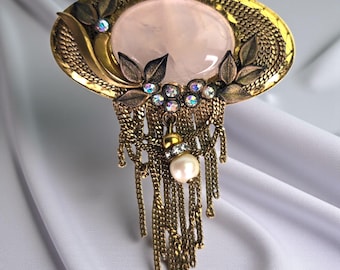 May include: An ornate, vintage-style brooch with a large oval, light pink stone at its center. The brooch features gold-toned metalwork, including leaves, chains, and sparkling crystals. A dangling pearl adds a touch of elegance.
