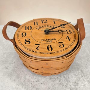 Könnte beinhalten: Eine runde Uhr mit Holzgesicht und schwarzen Ziffern sitzt auf einem geflochtenen Korb. Das Zifferblatt zeigt "Dresden Standard Time" und hat schwarze Zeiger. Ledergriffe sind an den Seiten der Uhr befestigt.