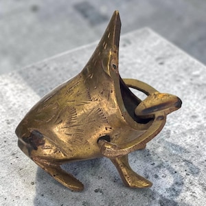 Pode incluir: Um abridor de garrafas vintage em forma de peixe de latão. O peixe tem a boca aberta com um abridor em forma de anel no interior. O latão apresenta detalhes gravados e uma pátina envelhecida. Objeto decorativo.