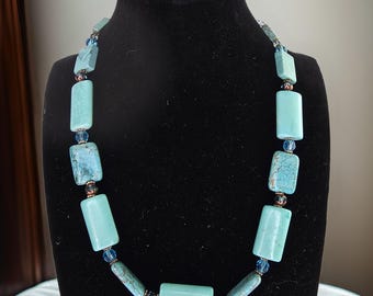 Op de afbeelding: Een turquoise en blauwe kralenketting met rechthoekige turquoise kralen en kleinere blauwe kralen. De ketting wordt getoond op een zwart manekin.
