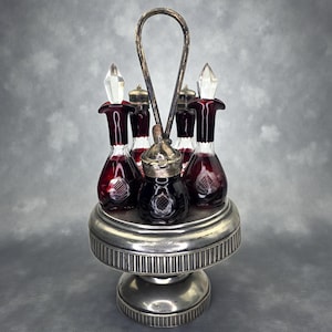 May include: An antique condiment set featuring six glass bottles with ruby red bodies and clear stoppers, set in a silver-toned metal carrier. The bottles sit on a tiered, pedestal base with a handle for easy transport. The set is designed for serving condiments.