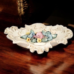 May include: An off-white, decorative ceramic dish with a scalloped edge and gold trim. The dish is adorned with three-dimensional, colorful flowers in shades of pink, blue, and yellow, creating a floral centerpiece.