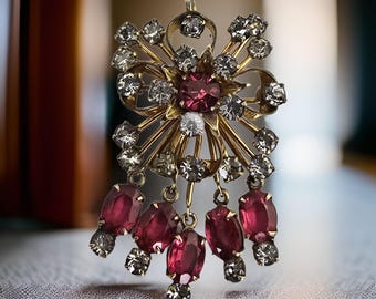 Puede incluir: Broche dorado vintage con diseño floral. El broche presenta una gran gema central de color rojo rubí rodeada de cristales transparentes más pequeños. Colgando del broche hay cinco piedras en forma de lágrima de color rojo rubí, cada una acentuada con un grupo de cristales transparentes.