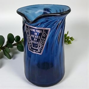 May include: A hand-blown, blue glass jug with a distinctive, wavy rim. The jug features a label stating "Hand Made in Mexico by Mexican" and "La Victoria". The glass has a textured, swirled pattern.