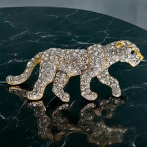 Puede incluir: Broche de leopardo enjoyado con contorno dorado y cristales transparentes. El broche está en posición de caminar, con la cabeza ligeramente girada a la derecha. El fondo es una superficie oscura y reflectante.