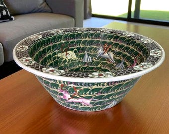 Könnte beinhalten: Eine dekorative Keramikschale mit breitem Rand und detailliertem Innendesign. Die Schale hat ein grün-weißes Muster mit Illustrationen von Figuren und Tieren. Der Rand hat ein kontrastierendes Muster aus Schwarz, Weiß und Rot. Die Schale steht auf einem Holztisch.