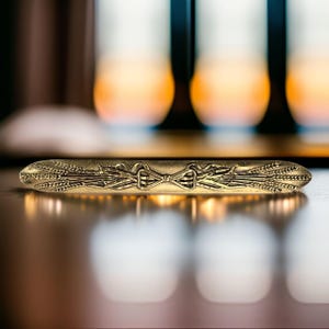 Könnte beinhalten: Ein goldfarbener Metall-Krawattenclip mit einem Weizenmuster. Der Krawattenclip liegt auf einer Holzoberfläche.