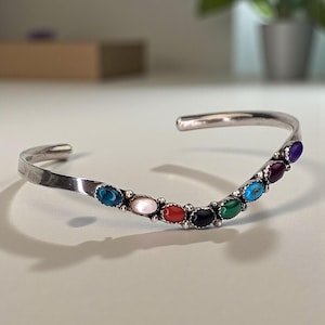 May include: A silver cuff bracelet with a row of colourful oval gemstones. Gemstones include turquoise, mother-of-pearl, red coral, onyx, and amethyst. A fashionable bracelet.