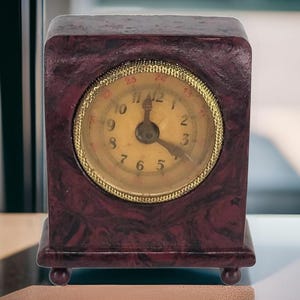 Może przedstawiać: Mały, kwadratowy zegar w kolorze bordowym z tarczą o złotej ramce i czarnymi wskazówkami. Tarcza zegara ma białe cyfry rzymskie i białe tło.