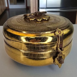 May include: A round, antique brass container with a lid. The lid features a floral design and a decorative handle. The container has a clasp closure and is likely used for storage. The brass has a warm, golden hue.