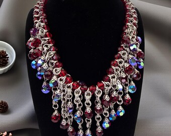 Puede incluir: Un collar llamativo con una cadena de cuentas rojas y eslabones de metal plateado. Cadenas colgantes de cuentas rojas y azules iridiscentes en forma de lágrima cuelgan de la cadena, creando un diseño glamuroso y llamativo.