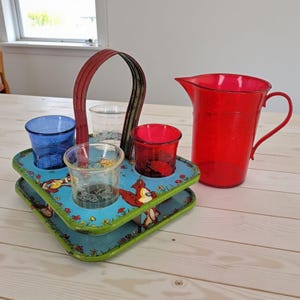 Puede incluir: Un juego de servir vintage con una jarra roja y una bandeja con cuatro vasos. La bandeja tiene un fondo azul con un diseño de dibujos animados y un asa roja. Los vasos son azules, rojos y transparentes.