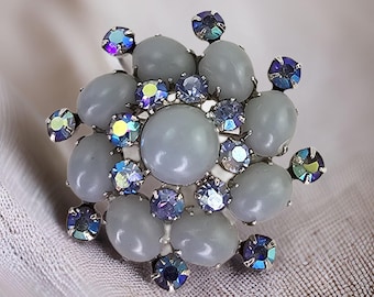 Puede incluir: Un broche de plata con un grupo de piedras cabujón grises y diamantes de imitación azules iridiscentes.