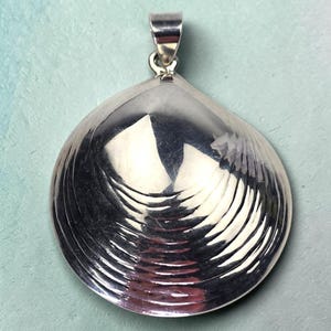 May include: A silver pendant in the shape of a clam shell. The pendant has a textured surface with curved lines and a loop at the top for a chain. The shell is highly reflective, with light reflecting off the surface.
