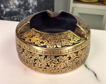Op de afbeelding: Ronde asbak met een zwart interieur en een goudkleurige buitenkant. De asbak heeft ingewikkelde bloemen- en bladerpatronen in goud, waardoor een decoratief en sierlijk ontwerp ontstaat. De asbak is gemaakt van metaal en heeft een klassieke vorm.