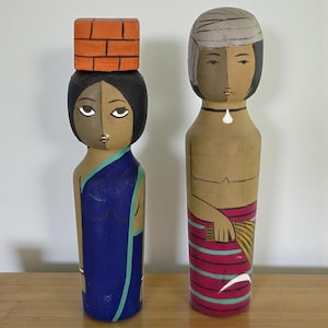 May include: Two wooden folk art figures. One figure is a woman in a blue sari with a brick-shaped object on her head. The other figure is a man with a turban and a striped skirt. Both figures have stylized facial features and are painted in earthy tones.