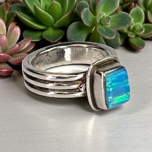 May include: A silver ring with a square, blue opal stone. The ring band has three horizontal grooves. The opal displays shades of blue and green. The ring is set against a blurred background of green succulents.