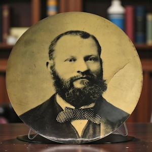 May include: A vintage portrait of a man with a full beard and a bow tie. The image is circular, with a sepia tone and some visible cracks. The man is wearing a dark suit. The portrait is displayed on a metal stand.