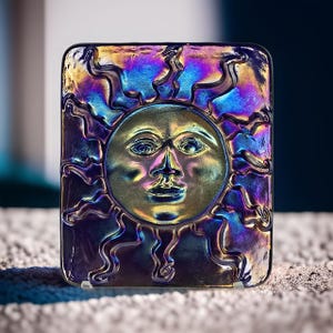 May include: A square piece of iridescent glass with a sun face design. The glass is a deep purple with blue and green hues. The sun face is embossed and has a gold colour.