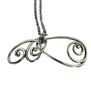 Może przedstawiać: Srebrny naszyjnik z wisiorkiem o abstrakcyjnym wzorze. Wisiorek ma wirujący, ażurowy wz&oacute;r z kwiatowym akcentem na g&oacute;rze, gdzie łączy się z łańcuszkiem. Łańcuszek jest delikatny, w stylu ogniw.