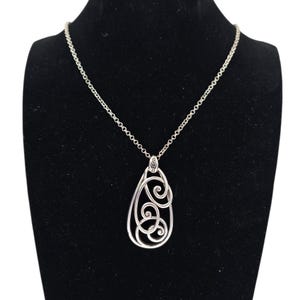 Może przedstawiać: Srebrny naszyjnik z wisiorkiem w kształcie łzy. Wisiorek ma ażurowy wz&oacute;r z wirującymi wzorami. Naszyjnik jest prezentowany na czarnej aksamitnej powierzchni.