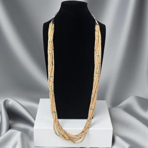 Puede incluir: Un collar de múltiples hilos con cuentas color beige y detalles turquesa. El collar se exhibe en un maniquí negro sobre un fondo de tela gris y blanca. Las cuentas están ensartadas, creando un efecto de capas.