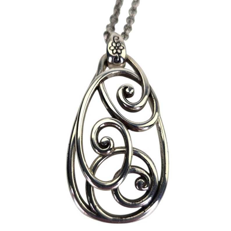 Może przedstawiać: Srebrny naszyjnik z wisiorkiem o otwartym kształcie łzy. Wisiorek ma wirujące, ażurowe wzory i jest zawieszony na delikatnym łańcuszku. G&oacute;rna część wisiorka ma kwiatowy wz&oacute;r.