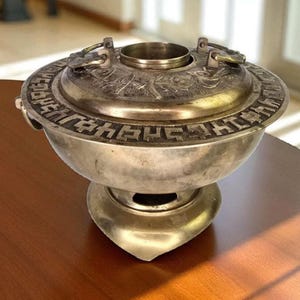 Puede incluir: Un cuenco de metal plateado con tapa con patrones y texto en relieve. El cuenco tiene una base redonda y una tapa decorativa con dos asas. El cuenco está sobre una superficie de madera.