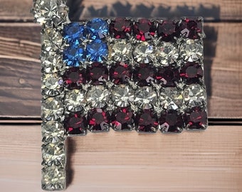May include: A rhinestone brooch shaped like an American flag. The flag is made of red, white and blue rhinestones. The brooch is a great way to show your patriotism.