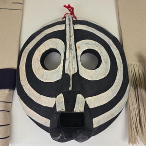 Könnte beinhalten: Eine schwarz-weiße Stammesmaske mit kreisförmigem Design. Die Maske hat zwei Augenlöcher und eine rechteckige Mundöffnung. Weiße Streifen umkreisen die Maske. Oben ist eine rote Schnur zum Aufhängen befestigt.