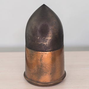 May include: A vintage artillery shell casing featuring a pointed, dark oxidized metal projectile tip attached to a polished, cylindrical copper base. The metal surface shows signs of wear, including scratches and natural patina throughout the object.