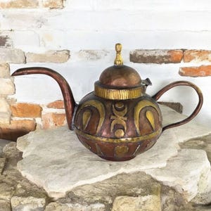 May include: A decorative metal teapot with a copper finish and gold accents. The teapot features a rounded body with ornate patterns, a curved spout, and a loop handle. The lid is topped with a small gold finial. The teapot is displayed on a stone surface.