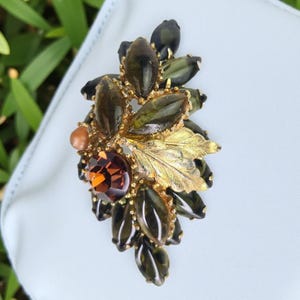 Könnte beinhalten: Eine Vintage-Brosche mit floralem Design. Die Brosche hat einen großen, runden, bernsteinfarbenen Edelstein in der Mitte, umgeben von olivgrünen und bernsteinfarbenen tropfenförmigen Steinen. Goldfarbene Blätter und ein kleiner, runder, pfirsichfarbener Stein vervollständigen das Design.
