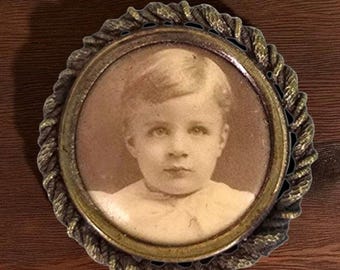 May include: A vintage sepia photograph of a young child, set within a decorative circular frame. The frame features a gold-toned inner rim and a textured, rope-style outer edge. The child has fair hair and wears a white top.