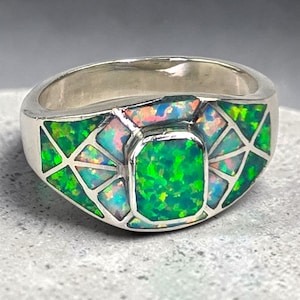 May include: A silver ring featuring a central rectangular opal inlay surrounded by a geometric pattern of smaller triangular and trapezoidal opal inlays. The opals display vibrant flashes of green, blue, and orange against a polished silver band.