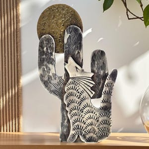 May include: A decorative metal sculpture of a cactus with a howling wolf silhouette. The sculpture features a textured finish and a brass-colored sun. The cactus is gray and white, with detailed etched patterns.