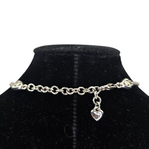 Może przedstawiać: Srebrny naszyjnik z wzorem łańcuszka. Naszyjnik posiada elementy dekoracyjne i mały urok w kształcie serca. Naszyjnik jest prezentowany na czarnym aksamitnym biuście.