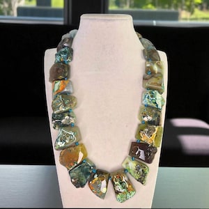 May include: A chunky necklace featuring large, irregularly shaped stones in shades of green, brown, and yellow. The stones are strung together with small blue beads. The necklace is displayed on a white mannequin.