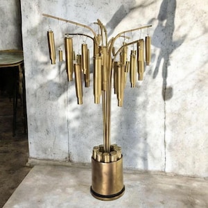 Op de afbeelding: Een messingkleurige decoratieve sculptuur in de vorm van een boom. De sculptuur heeft een centrale stam met meerdere takken, waaraan cilindrische elementen hangen. De basis is een cilindrische container gevuld met vergelijkbare elementen.