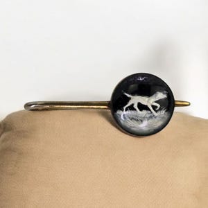 May include: A vintage hair barrette with a round, black enamel design featuring a white dog silhouette running through grass. The barrette has a gold-toned metal stem and a simple, elegant design.