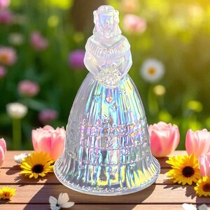 May include: A clear, iridescent glass figurine of a woman in a long dress, holding a bouquet. The figurine is set on a wooden surface with pink tulips and yellow daisies in the background. The dress has a detailed, textured design.